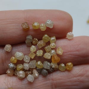 May include: A collection of small, uncut diamonds in various shades of yellow, brown, and clear. The rough gemstones are held in a hand, showcasing their natural, irregular shapes and sparkling facets. The diamonds vary in size and clarity.