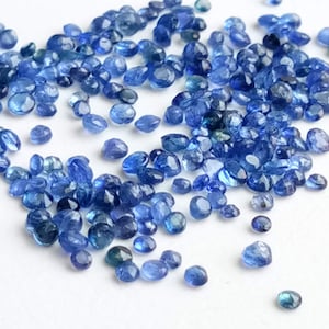 May include: A loose pile of small, faceted, blue gemstones. The gemstones are scattered on a white surface.
