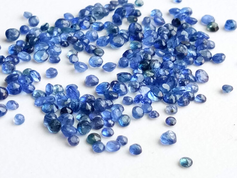 May include: A loose pile of small, faceted, blue gemstones. The gemstones are scattered on a white surface.