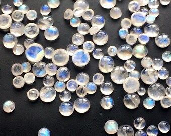Puede incluir: Una colección de cabujones redondos de piedra lunar iridiscente de varios tamaños. Las piedras son de color blanco lechoso con un brillo azul.