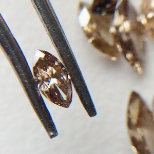 May include: A close-up of a light brown marquise-shaped diamond held by tweezers. The diamond has a slight golden hue and is surrounded by other loose diamonds.