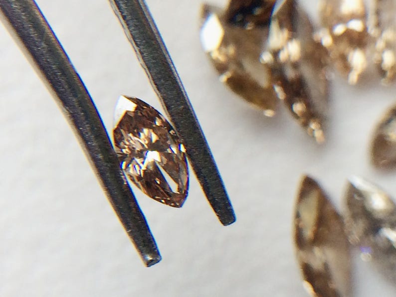 May include: A close-up of a light brown marquise-shaped diamond held by tweezers. The diamond has a slight golden hue and is surrounded by other loose diamonds.