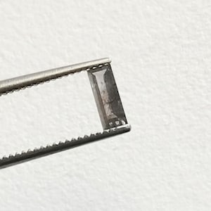 May include: A rectangular, gray gemstone is held by a pair of silver tweezers. The gemstone has a dark, smoky appearance with visible internal features. The tweezers are in sharp focus against a plain white background.