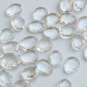 Peut inclure: Une collection de cristaux de quartz clairs, facet&eacute;s et de forme ovale. Les cristaux sont dispers&eacute;s sur une surface blanche.