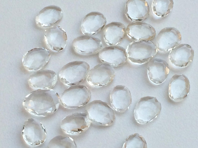 Peut inclure: Une collection de cristaux de quartz clairs, facet&eacute;s et de forme ovale. Les cristaux sont dispers&eacute;s sur une surface blanche.