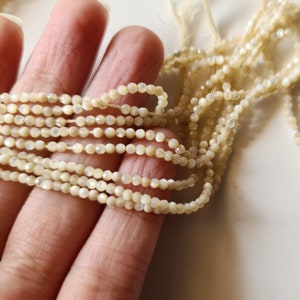 K&ouml;nnte beinhalten: Eine Str&auml;hne aus wei&szlig;en, irisierenden Muschelkugeln, etwa 4 mm im Durchmesser, auf einem d&uuml;nnen wei&szlig;en Faden aufgereiht. Die Perlen haben eine facettierte, leicht unregelm&auml;&szlig;ige Form.