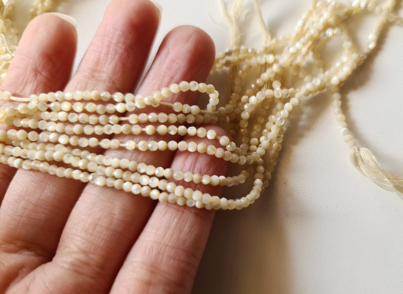K&ouml;nnte beinhalten: Eine Str&auml;hne aus wei&szlig;en, irisierenden Muschelkugeln, etwa 4 mm im Durchmesser, auf einem d&uuml;nnen wei&szlig;en Faden aufgereiht. Die Perlen haben eine facettierte, leicht unregelm&auml;&szlig;ige Form.