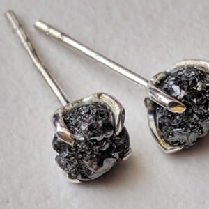 May include: A pair of silver stud earrings with black rough diamonds. The diamonds are set in a prong setting and the earrings have a post and butterfly back.