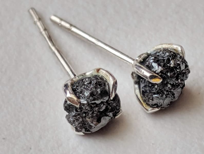 May include: A pair of silver stud earrings with black rough diamonds. The diamonds are set in a prong setting and the earrings have a post and butterfly back.