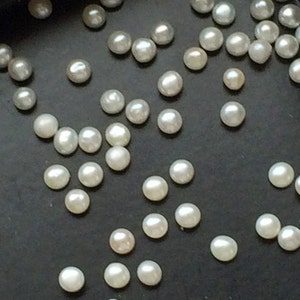 May include: A collection of white pearls scattered on a black surface. The pearls are of various sizes and shapes.