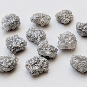 K&ouml;nnte beinhalten: Eine Sammlung von zehn rohen, grauen Diamanten. Die Diamanten haben verschiedene Formen und Gr&ouml;&szlig;en und haben ein nat&uuml;rliches, unpoliertes Aussehen.