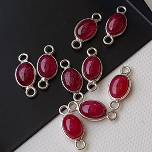 May include: A group of eight oval-shaped red gemstone connector charms with silver-toned metal loops. The charms are scattered on a black and white surface.