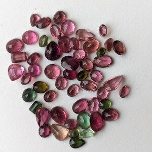 May include: A collection of various-shaped gemstones in shades of pink, green, and clear. The gemstones are scattered across a white surface. The shapes include oval, round, pear, and rectangular cuts. The gemstones vary in size.