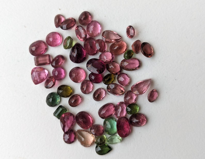 May include: A collection of various-shaped gemstones in shades of pink, green, and clear. The gemstones are scattered across a white surface. The shapes include oval, round, pear, and rectangular cuts. The gemstones vary in size.