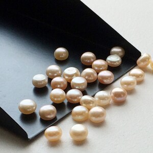 May include: A collection of round, off-white pearls with a slight pink hue. The pearls are scattered on a black surface.