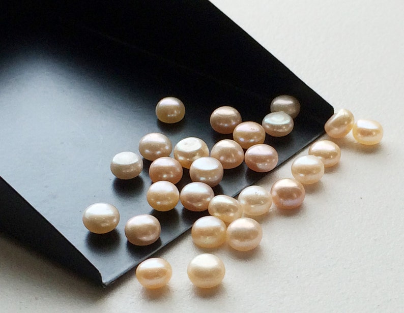 May include: A collection of round, off-white pearls with a slight pink hue. The pearls are scattered on a black surface.