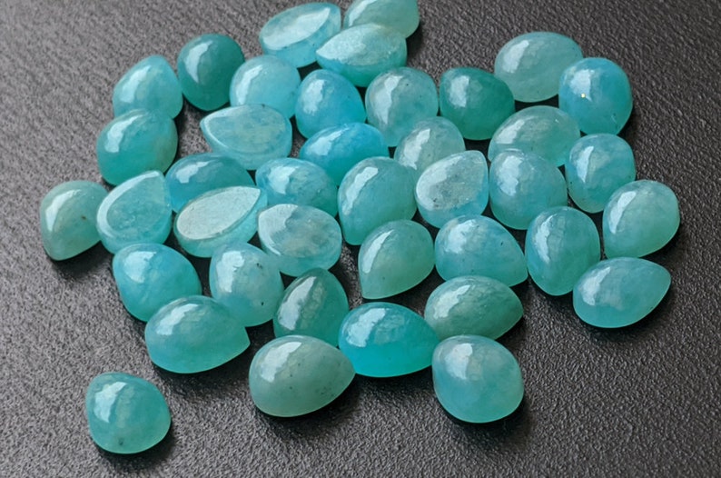 May include: A collection of numerous loose, teardrop-shaped gemstones in a vibrant turquoise-blue color. Each stone features a smooth, polished, and rounded cabochon surface that reflects light, showcasing a consistent semi-translucent appearance.