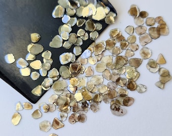 May include: A collection of small, irregularly shaped, translucent, light brown and green gemstones. The gemstones are scattered on a white surface.