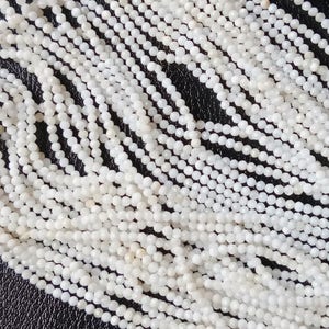 May include: A strand of white gemstone beads, each bead is round and has a slightly faceted surface. The beads are strung together on a clear thread.