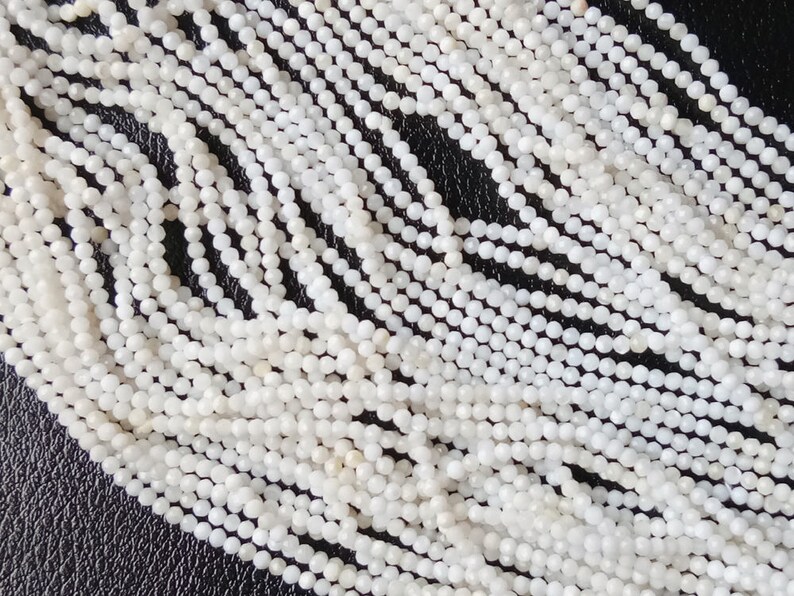 May include: A strand of white gemstone beads, each bead is round and has a slightly faceted surface. The beads are strung together on a clear thread.
