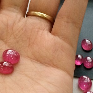 Pu&ograve; includere: Primo piano di una mano che tiene due gemme rosa. Le gemme sono rotonde e hanno una superficie liscia e lucida. Ci sono diverse altre gemme rosa su una superficie grigia sullo sfondo.