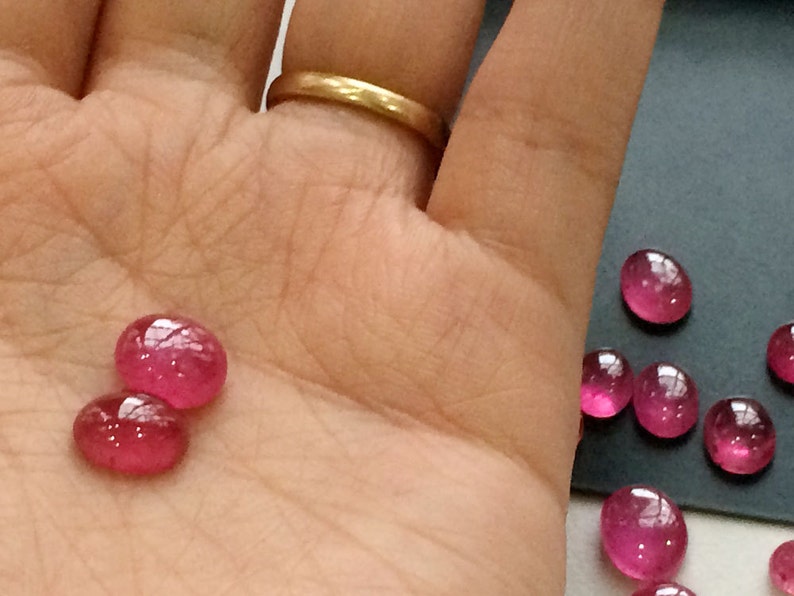 Pu&ograve; includere: Primo piano di una mano che tiene due gemme rosa. Le gemme sono rotonde e hanno una superficie liscia e lucida. Ci sono diverse altre gemme rosa su una superficie grigia sullo sfondo.