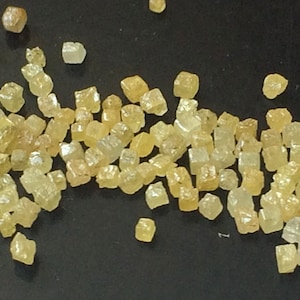 May include: A close-up of a collection of small, yellow, faceted, cubic crystals. The crystals are scattered on a black surface.