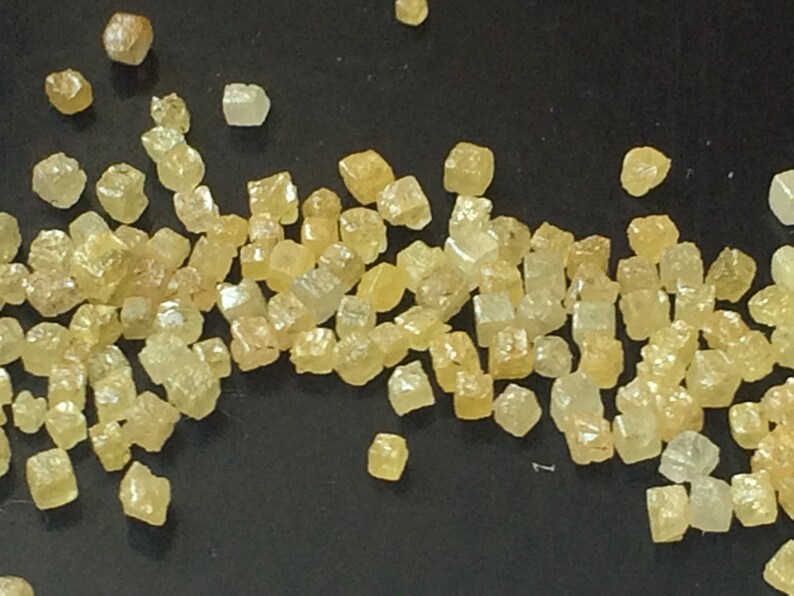 May include: A close-up of a collection of small, yellow, faceted, cubic crystals. The crystals are scattered on a black surface.