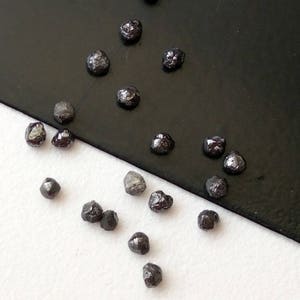 May include: A collection of black diamonds, some with a slight grey hue, scattered on a white and black surface. The diamonds are various sizes and shapes.