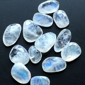May include: A collection of 12 smooth, white moonstone gemstones with iridescent blue and white flashes. The stones are faceted and have a slightly oval shape.