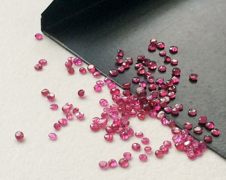 May include: A scattering of small, faceted, ruby-red gemstones. The gems are scattered across a surface, with some appearing to be in a small, black tray. The gems have a brilliant, sparkling appearance, suggesting they are of high quality.