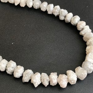 Op de afbeelding: Een streng ruwe, witte, natuurlijke diamanten die als ketting aan elkaar geregen zijn. De diamanten zijn onregelmatig van vorm en hebben een matte afwerking.