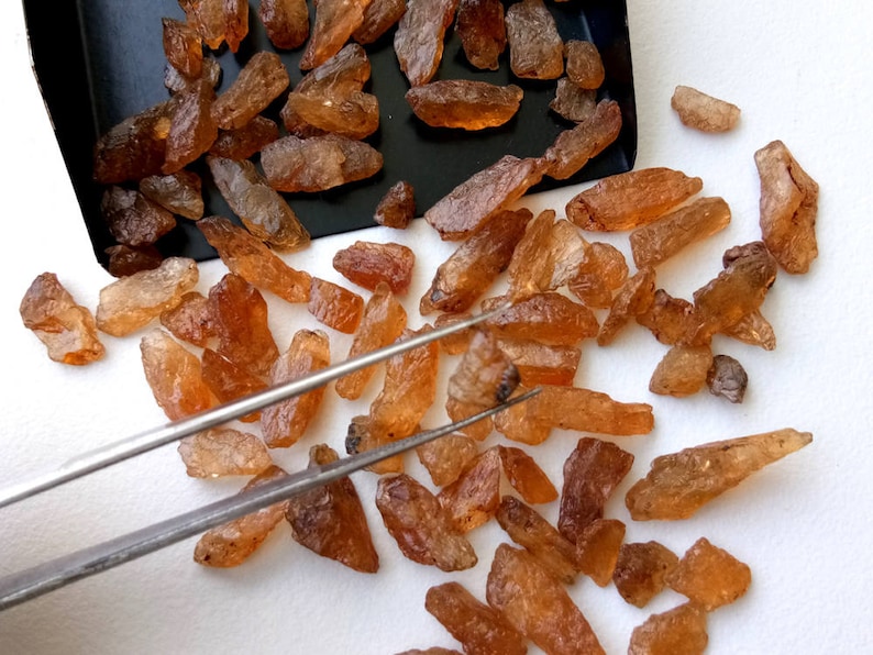 May include: A collection of small, rough, brown gemstones, some with a reddish hue, scattered on a white surface. The gemstones are arranged in a random pattern, with some overlapping. A pair of tweezers is visible in the image, suggesting that the gemstones are being sorted or examined.
