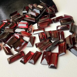May include: A pile of loose red gemstone rectangular facets. The facets are cut and polished with a high shine.