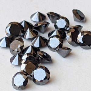 May include: A collection of black round faceted gemstones, likely diamonds, scattered on a white surface. The gemstones are various sizes and have a shiny, reflective surface.