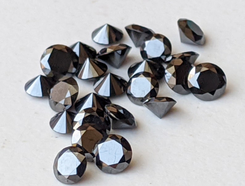 May include: A collection of black round faceted gemstones, likely diamonds, scattered on a white surface. The gemstones are various sizes and have a shiny, reflective surface.
