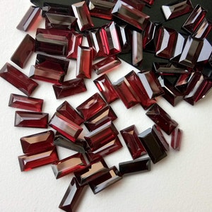 May include: A collection of deep red rectangular gemstones, likely garnets, scattered on a white surface. The gemstones are faceted and have a glossy finish.