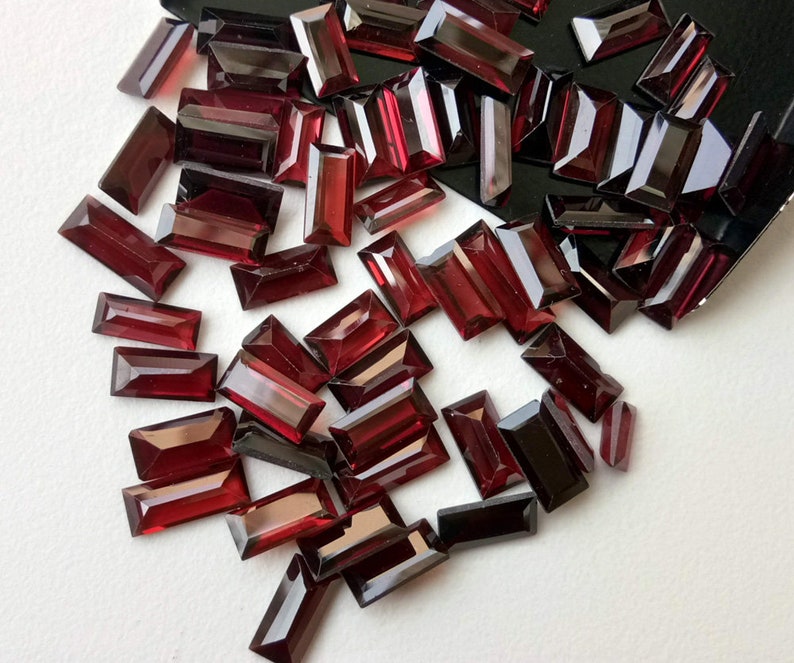 May include: A collection of deep red rectangular gemstones, likely garnets, scattered on a white surface. The gemstones are faceted and have a glossy finish.