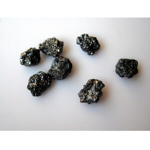 May include: A collection of seven black diamonds, also known as carbonado diamonds, on a white surface. The diamonds are rough and unpolished, with a variety of shapes and sizes.