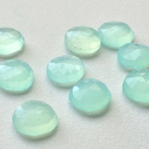 May include: A collection of nine round, faceted gemstones in a pale, translucent aqua color. The gemstones are scattered on a white surface, each displaying a subtle, internal glow and multiple facets.