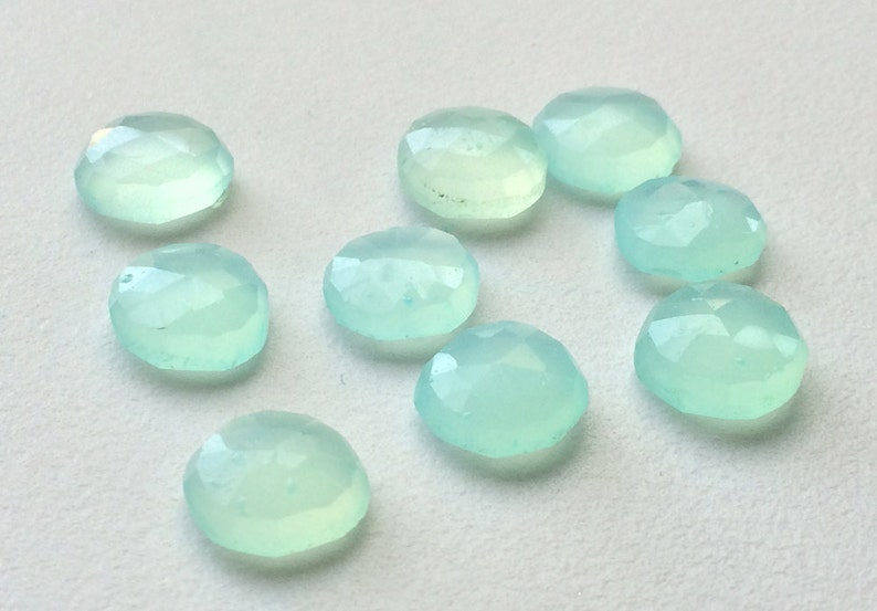 May include: A collection of nine round, faceted gemstones in a pale, translucent aqua color. The gemstones are scattered on a white surface, each displaying a subtle, internal glow and multiple facets.