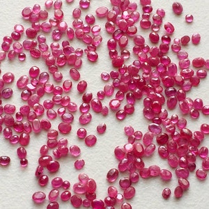 May include: A large group of loose, oval-shaped, pink ruby gemstones. The rubies are scattered on a white surface.