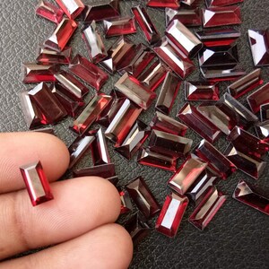 May include: A large quantity of dark red, rectangular-shaped glass rhinestones. The rhinestones are scattered on a black surface. One rhinestone is being held by a finger.