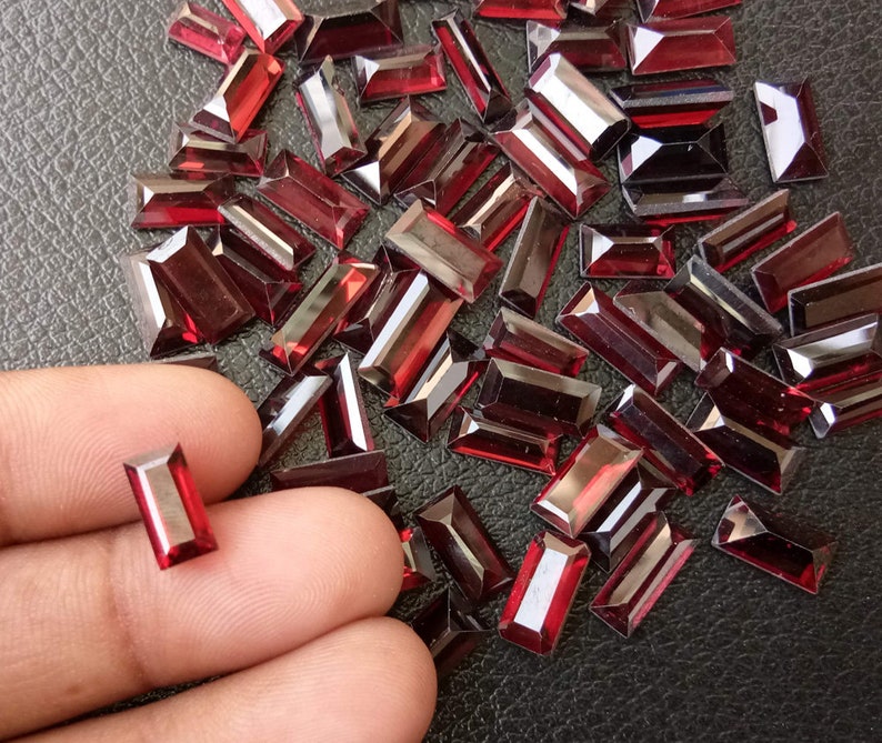 May include: A large quantity of dark red, rectangular-shaped glass rhinestones. The rhinestones are scattered on a black surface. One rhinestone is being held by a finger.