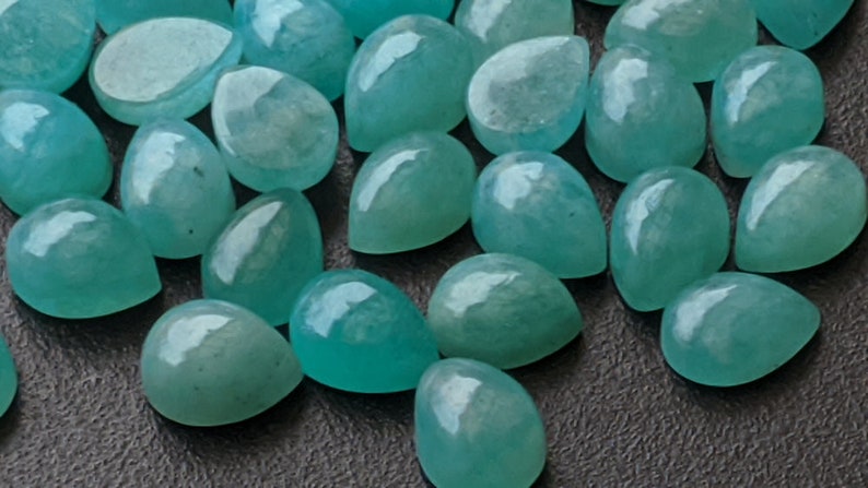 May include: A collection of loose, teardrop-shaped gemstones in a vibrant turquoise-blue color. Each stone features a smooth, polished, cabochon-style surface with a glossy finish, reflecting light across their rounded, symmetrical forms.