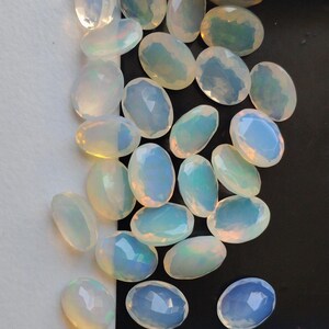 May include: A collection of oval-shaped, faceted opals. The opals display a milky white base color with iridescent flashes of blue, green, and yellow. The gemstones are arranged on a black and white background.