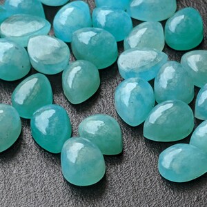 May include: A collection of numerous loose, teardrop-shaped gemstones in a vibrant turquoise-blue color. Each stone features a smooth, polished, domed cabochon surface that reflects light, showcasing varying degrees of translucency and internal texture.