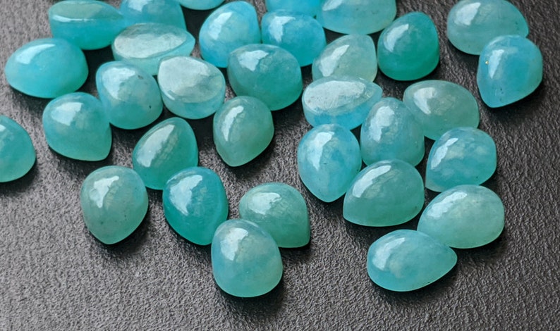 May include: A collection of numerous loose, teardrop-shaped gemstones in a vibrant turquoise-blue color. Each stone features a smooth, polished, domed cabochon surface that reflects light, showcasing varying degrees of translucency and internal texture.