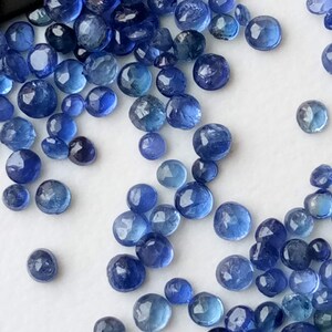 May include: A collection of loose blue gemstone beads, scattered on a white surface. The beads are round and have a slightly faceted appearance.