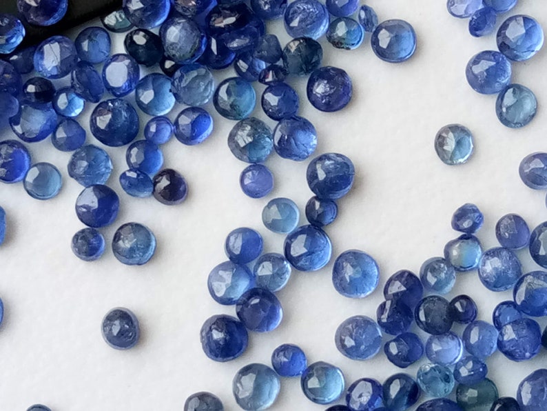 May include: A collection of loose blue gemstone beads, scattered on a white surface. The beads are round and have a slightly faceted appearance.